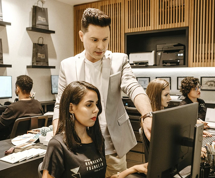 Fotografía de varias personas usando una computadora, y un hombre dándole instrucciones a una chica.