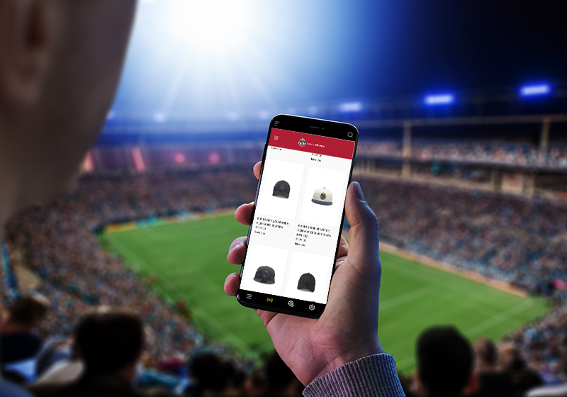 Fotografía de un aficionado de football en la cancha viendo la tienda online de su equipo en el celular.